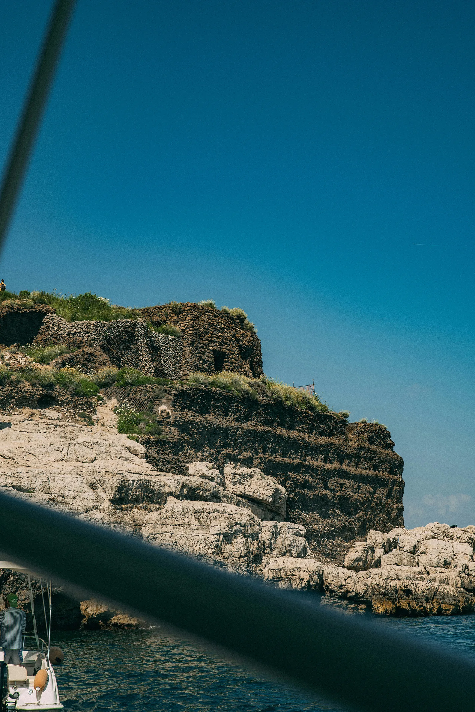 Rocky coastal cliff with green vegetation rising above clear blue sea under a bright sky.
