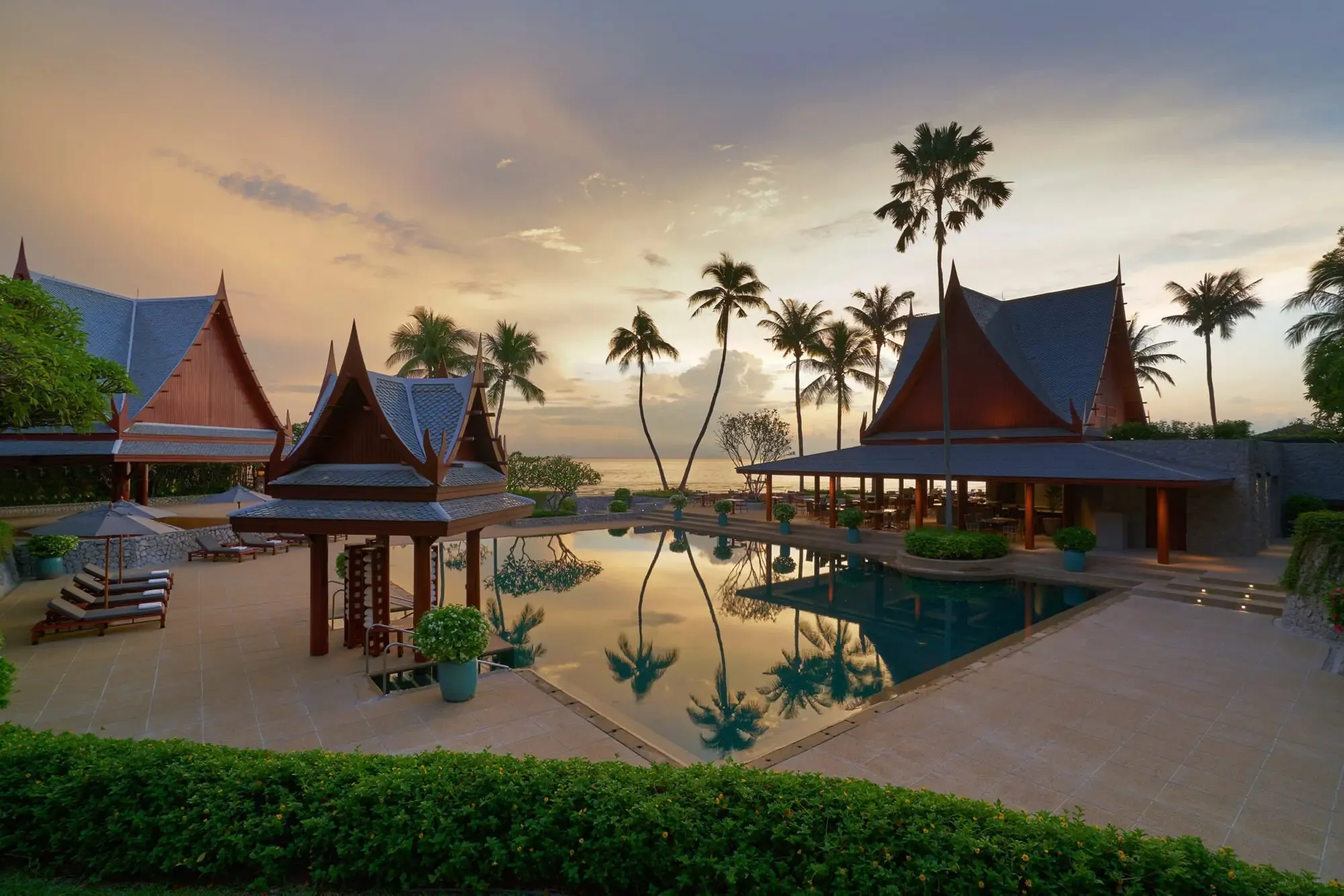 Outdoor pool at Chiva Som surrounded by traditional Thai pavilions and palm trees at sunset.