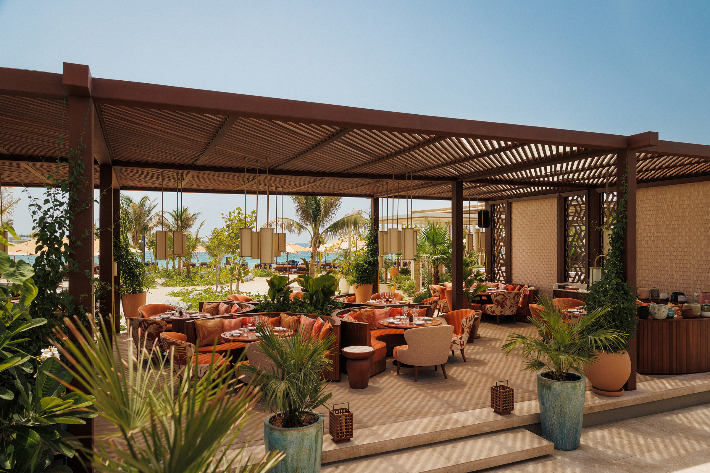 An outdoor dining venue with orange seating and plants shaded by a wooden canopy beneath a clear sky
