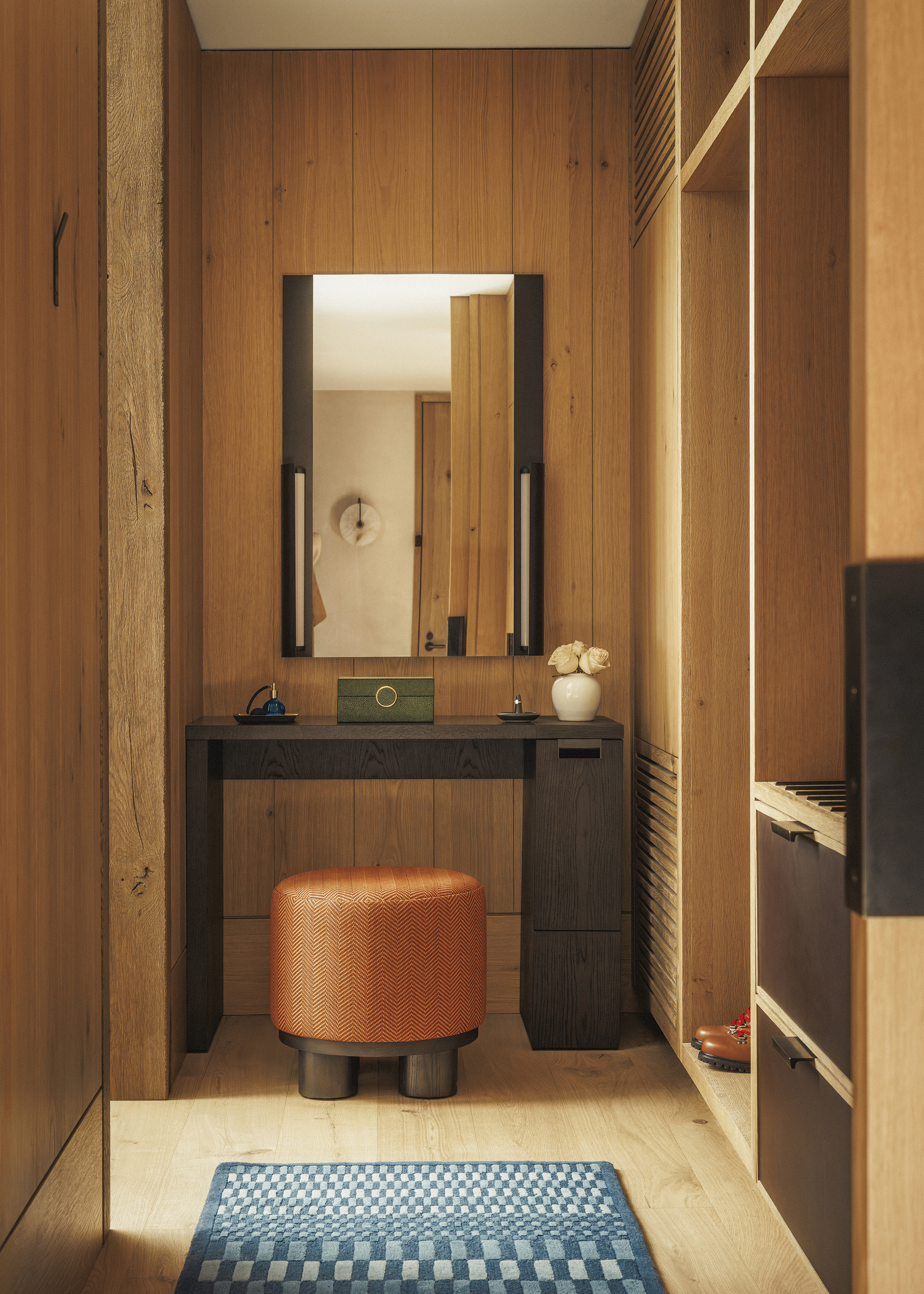 A cosy vanity area featuring a large mirror and table with decorative items above a round orange stool surrounded by wooden wardrobes