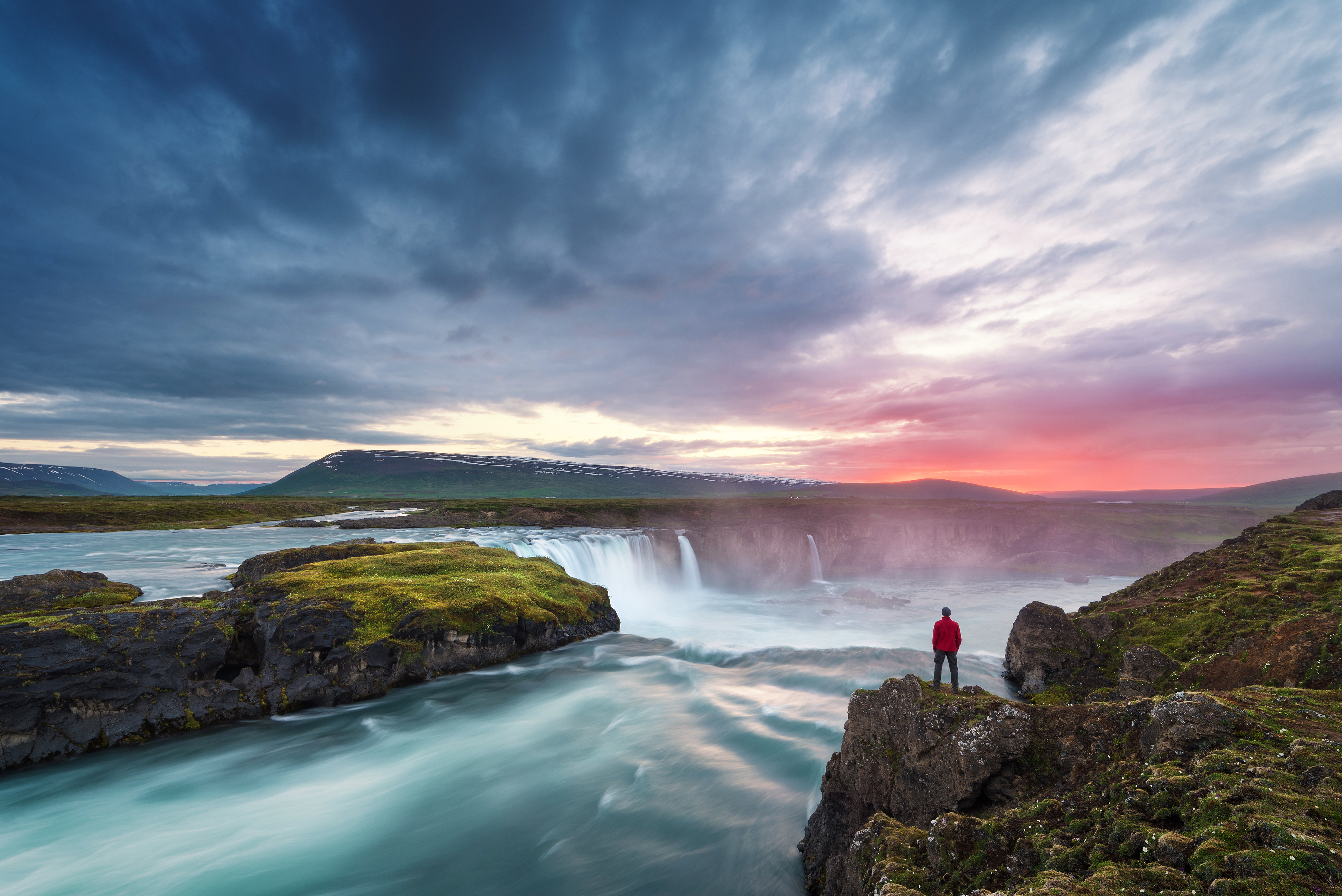 A man in a red coat standing among a vast landscape on the edge of Goðafoss Waterfall