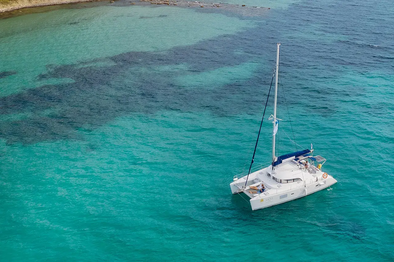 White catamaran anchored in clear turquoise water near a rocky coastline on a sunny day.