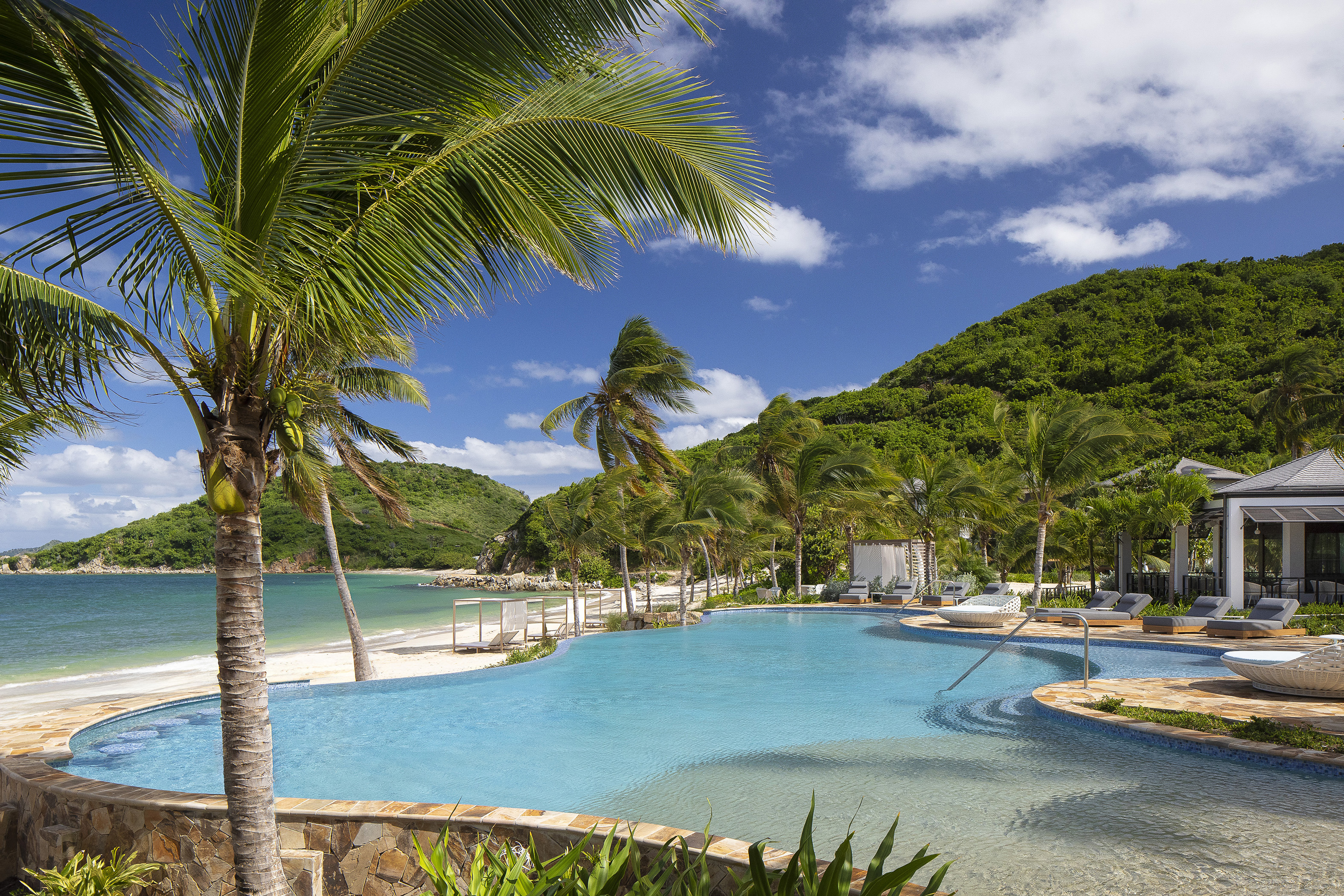 Blue infinity pool flanked by palm trees in front of lush green hills with a white pavilion at the side