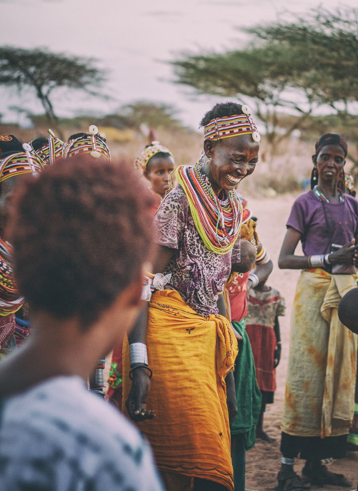 A group of people in traditional colorful African clothing and jewelry stand outdoors with trees in the background.