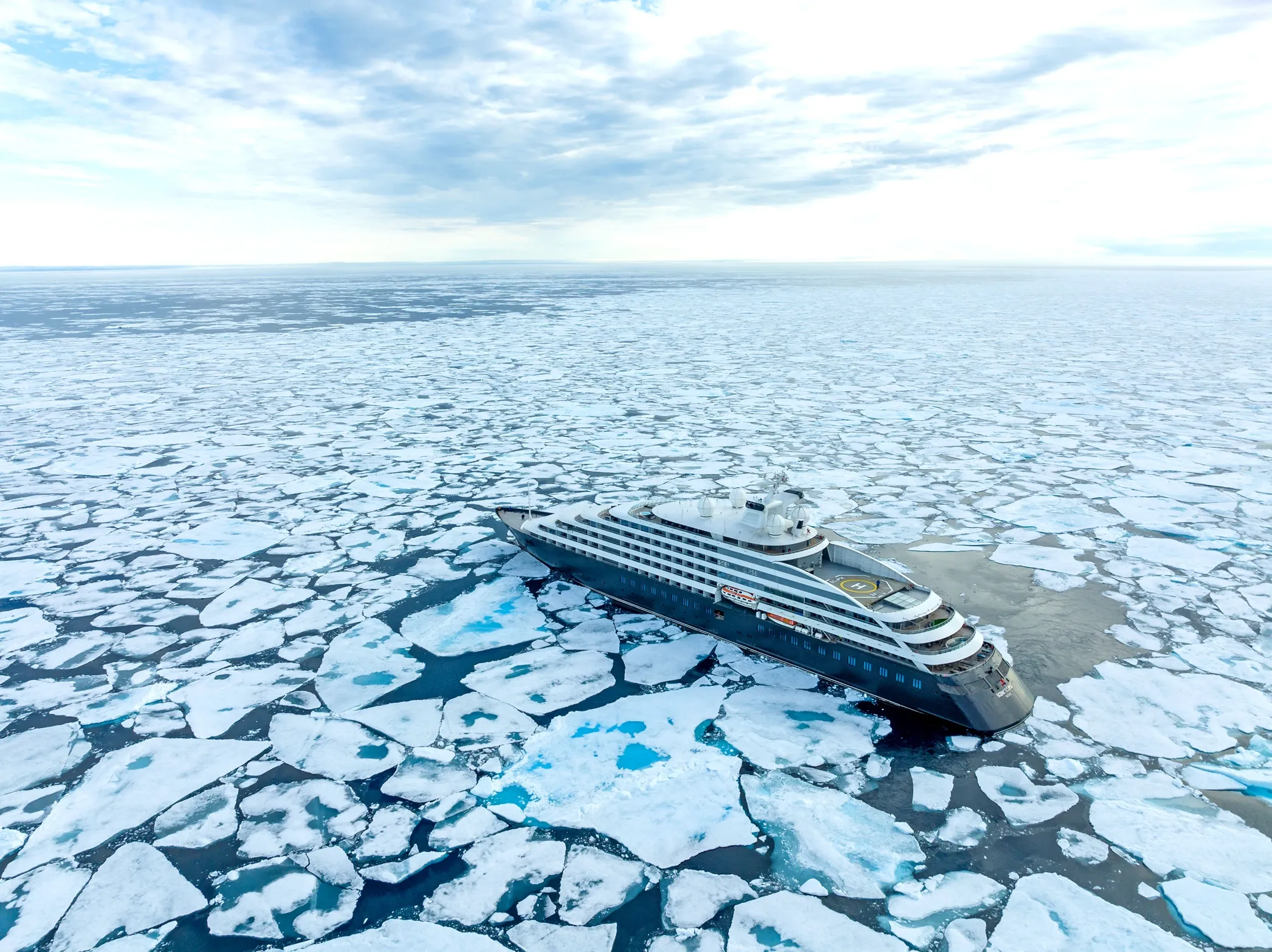 Cruise ship sailing through floating ice in polar waters under a cloudy sky.