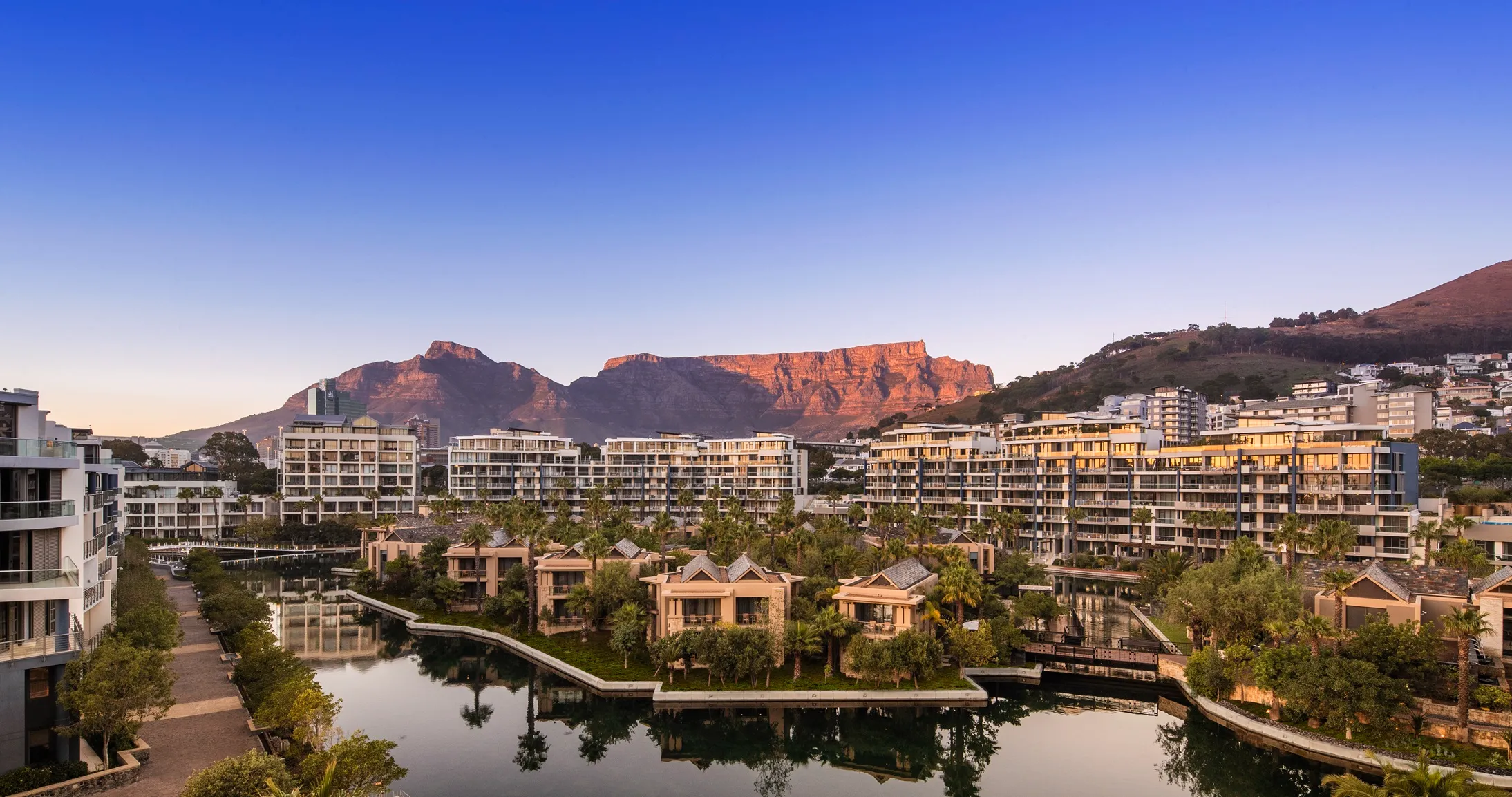 A scenic view of One&Only Cape Town, with modern buildings, lush greenery, and a water body in the foreground, set against mountains under a clear blue sky.
