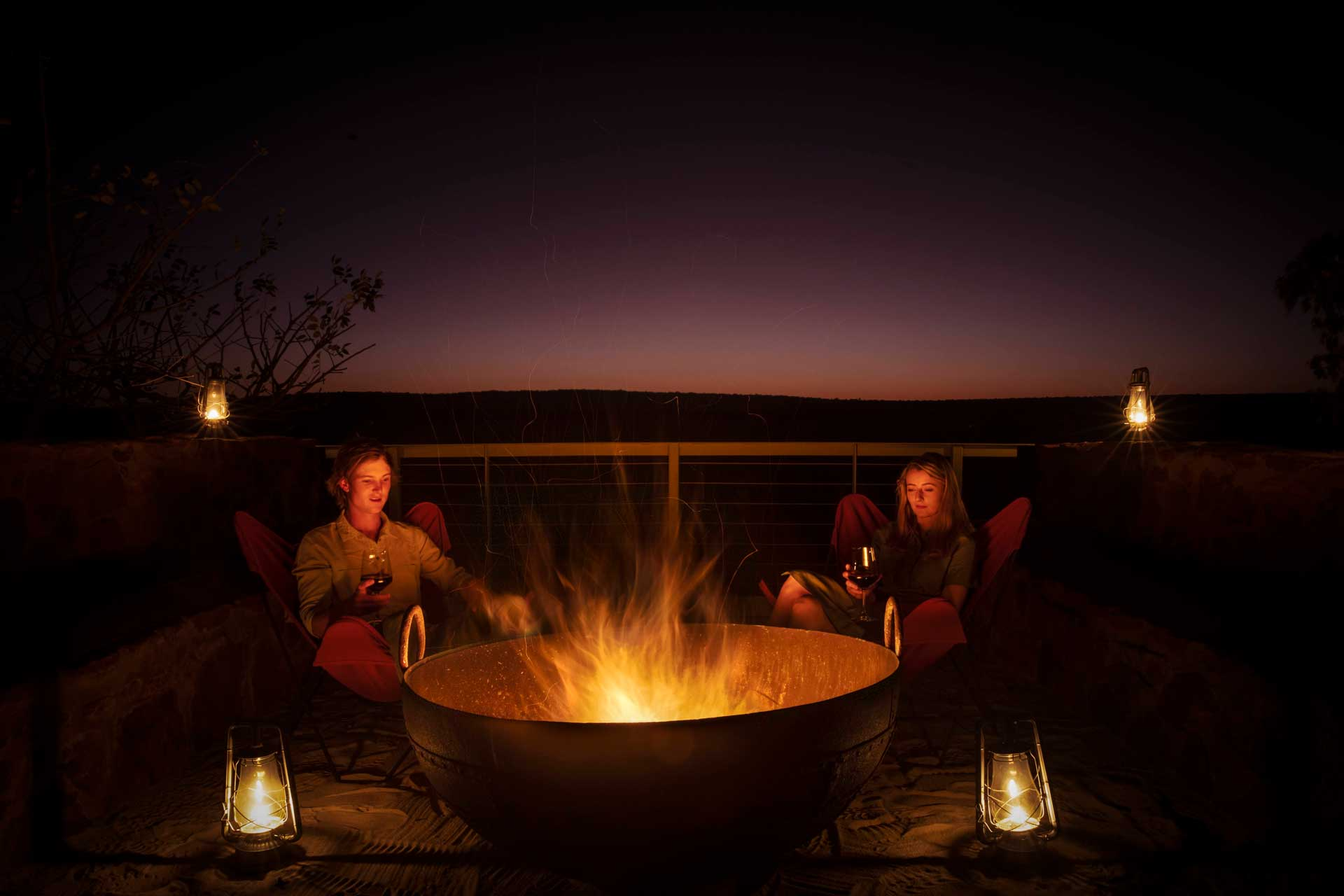 Two guests sitting by a fire pit at night, illuminated by lanterns while enjoying drinks under a dark sky.