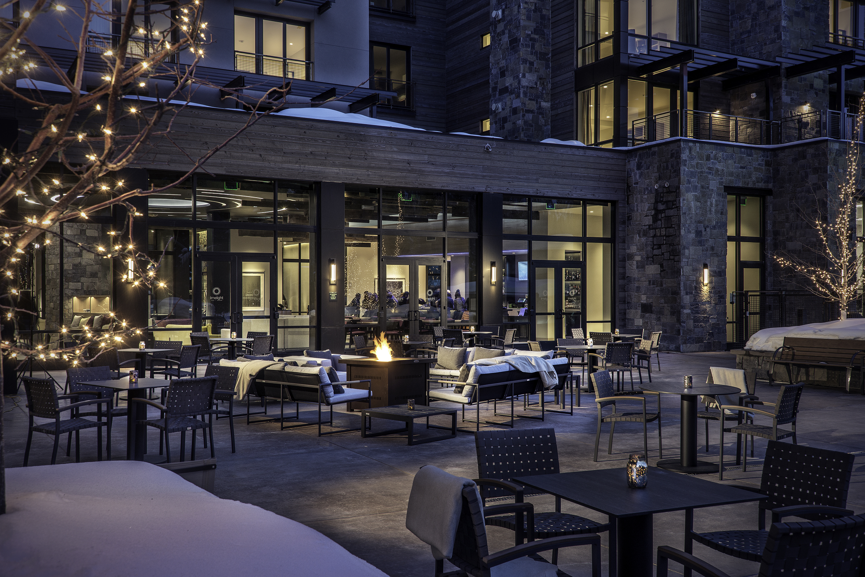 Outdoor patio area at Limelight Ketchum hotel featuring tables, chairs, a fire pit, snow-covered ground, and illuminated trees