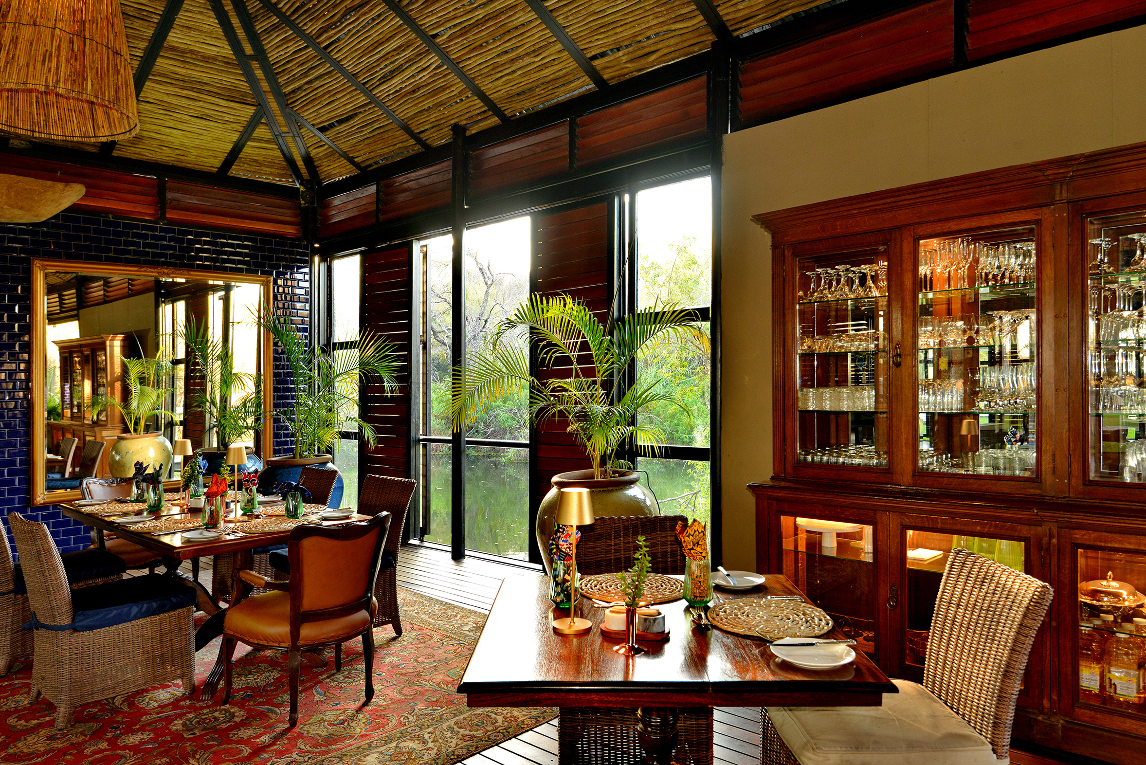 An inviting dining and bar space at Mukwa River Lodge featuring a thatched ceiling, lush plants, elegant furnishings, and warm natural light