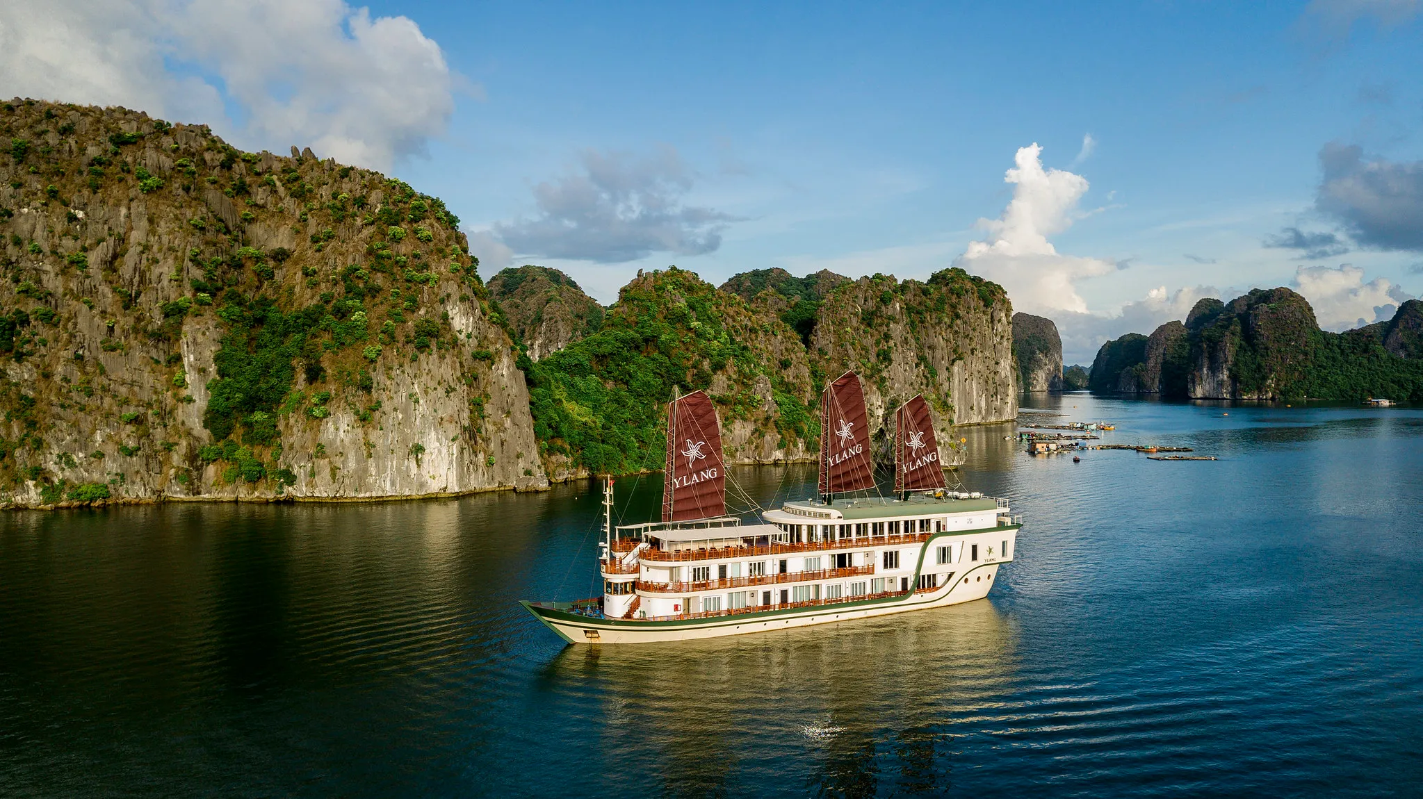Heritage Line boat with red sails cruising through a bay of limestone cliffs and lush greenery under a partly cloudy sky.