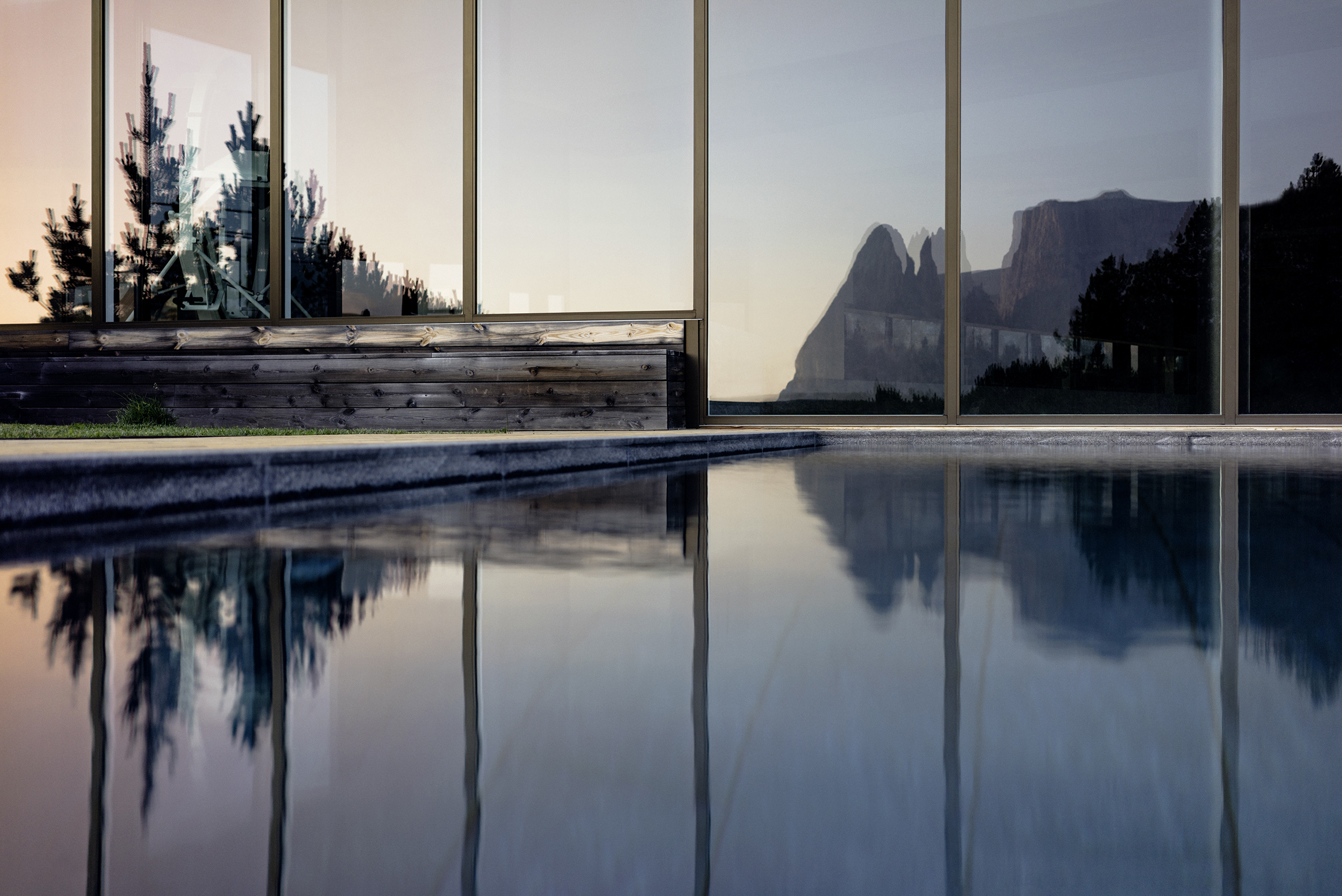 A tranquil outdoor pool reflecting the silhouette of a mountain range at dusk, viewed through large glass windows that also reflect an interior space with plants.