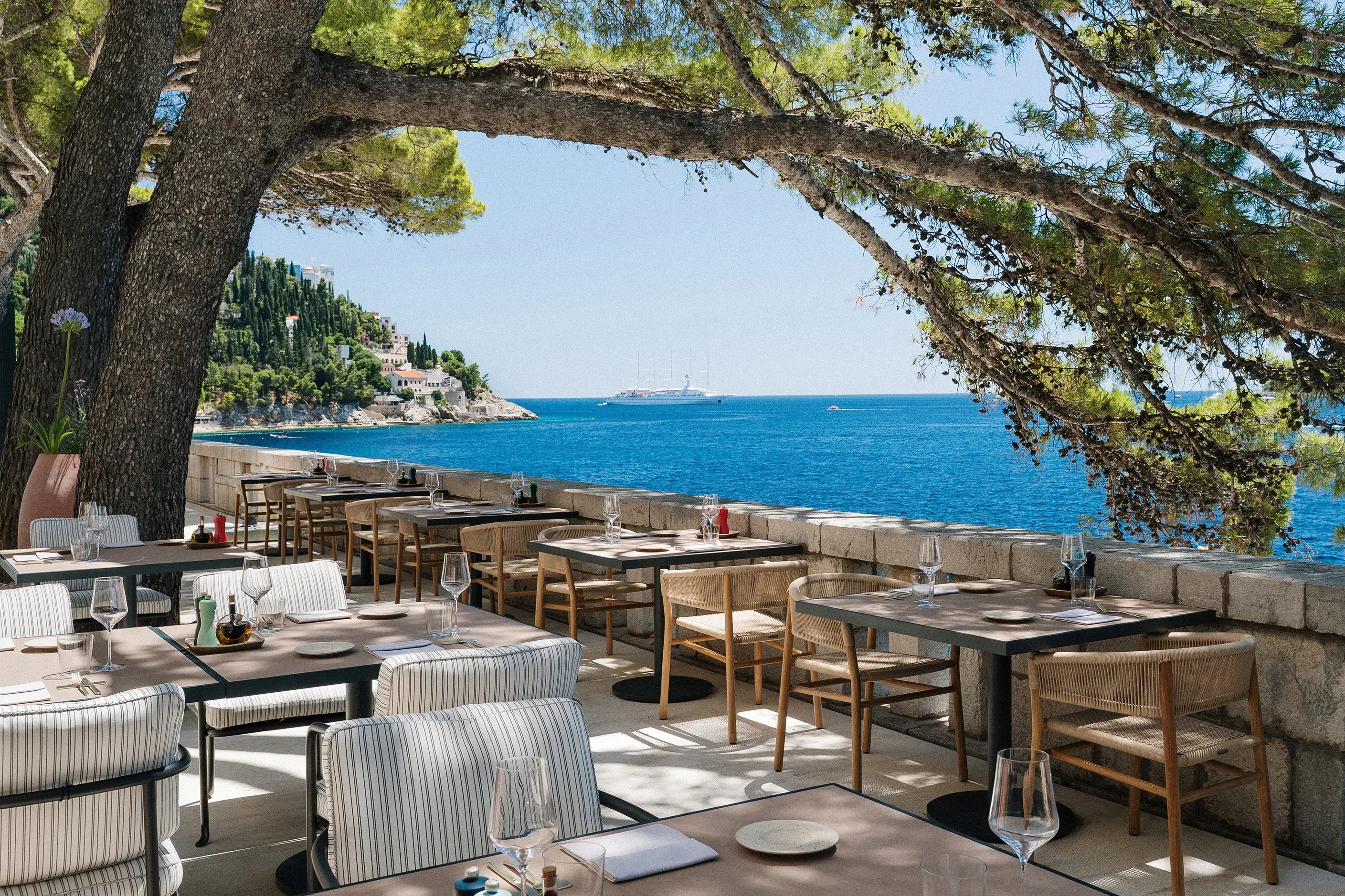 Giardino Restaurant al fresco terrace at Villa Dubrovnik in Croatia featuring dining tables and chairs set beneath pine trees beside the sea with a view of a yacht behind