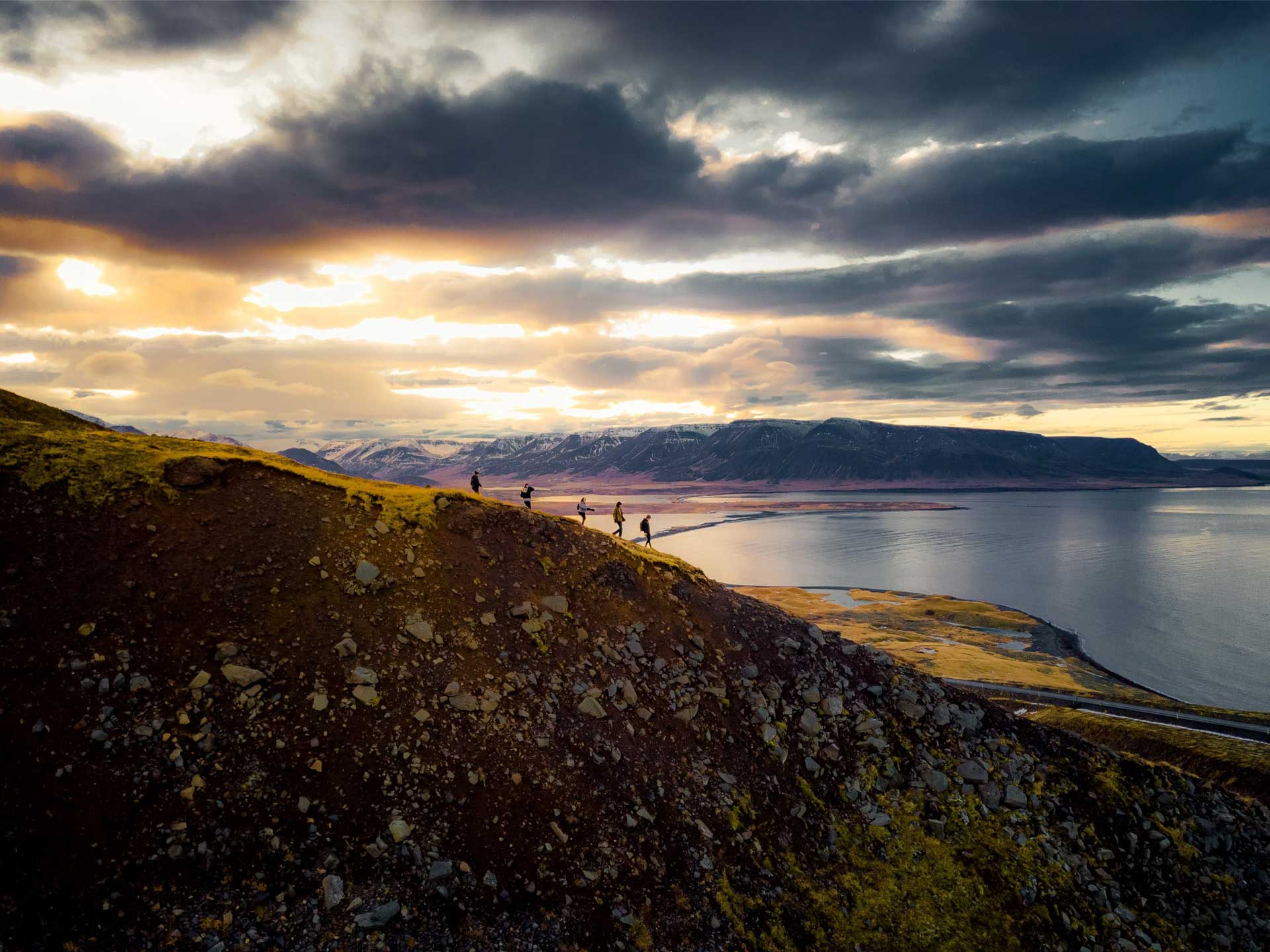 Group of hikers walking along a rugged coastal ridge at sunset with expansive mountain and sea views.