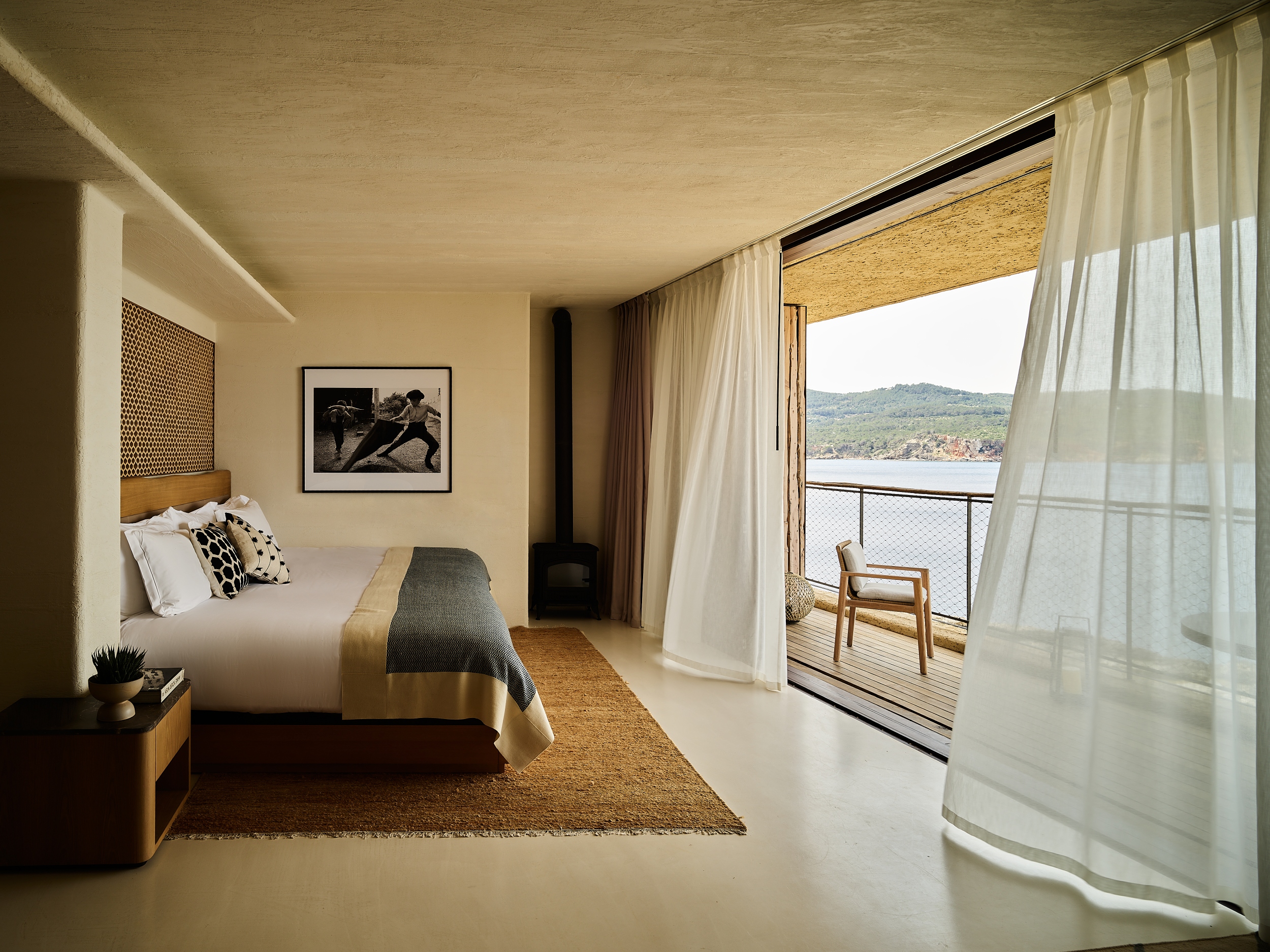 Modern bedroom with a large bed, photograph, and balcony overlooking a lake and hills.