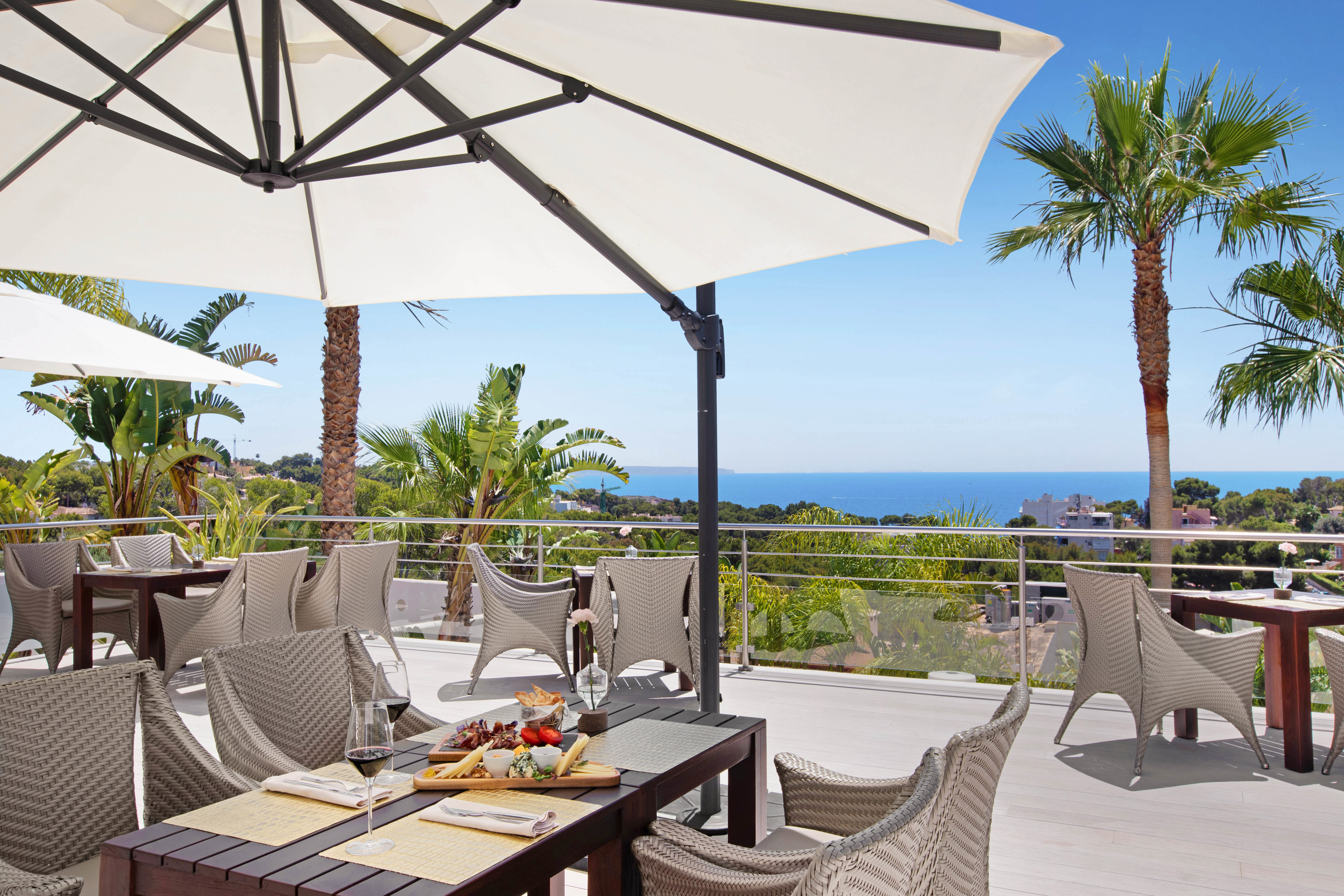 Outdoor dining area at Portals Hills in Mallorca with wicker chairs, white umbrellas, palm trees, and sea views.