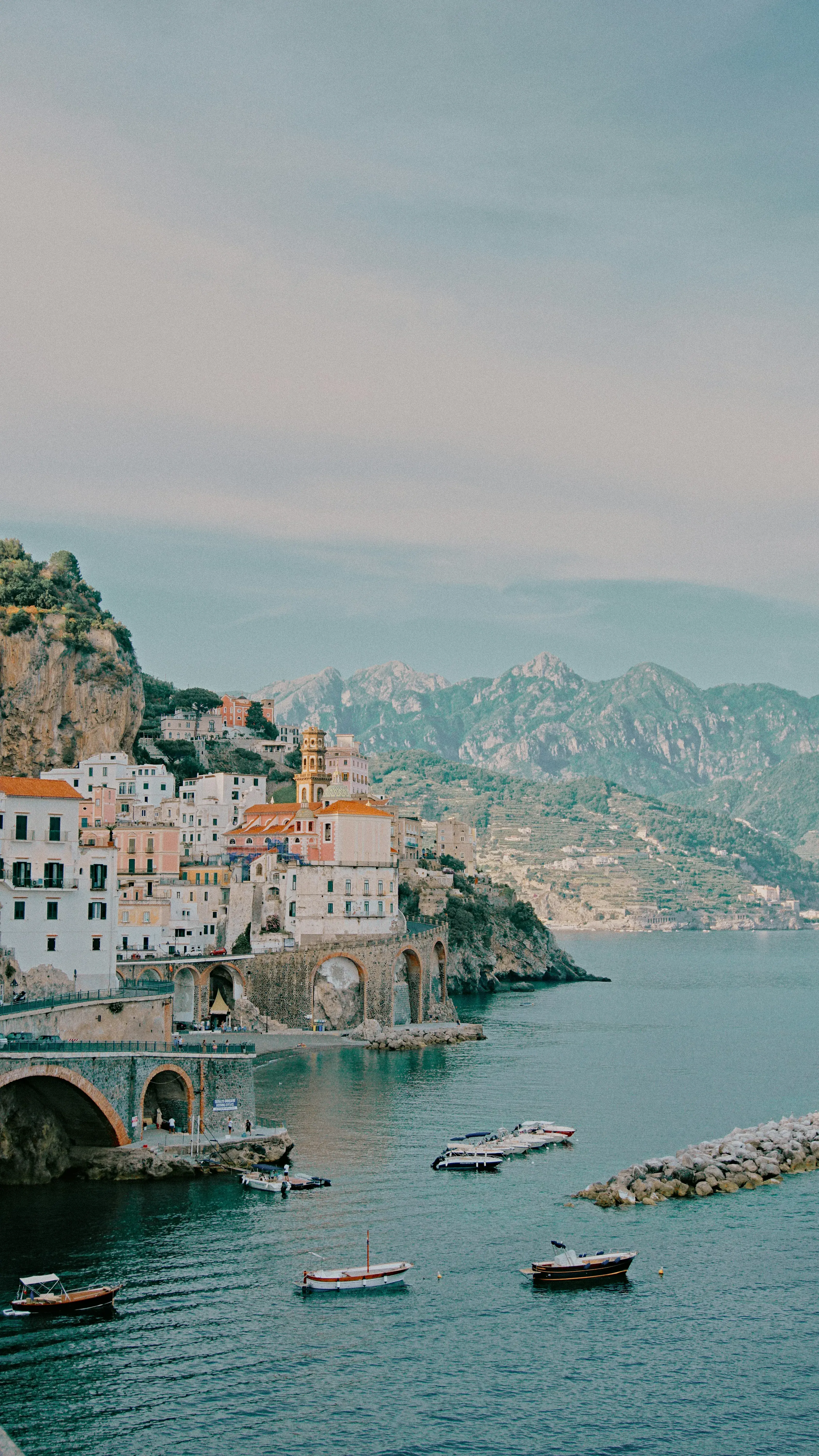 Scenic view of Amalfi Coast with colorful seaside buildings, arched bridge, and boats on turquoise water against mountain backdrop.