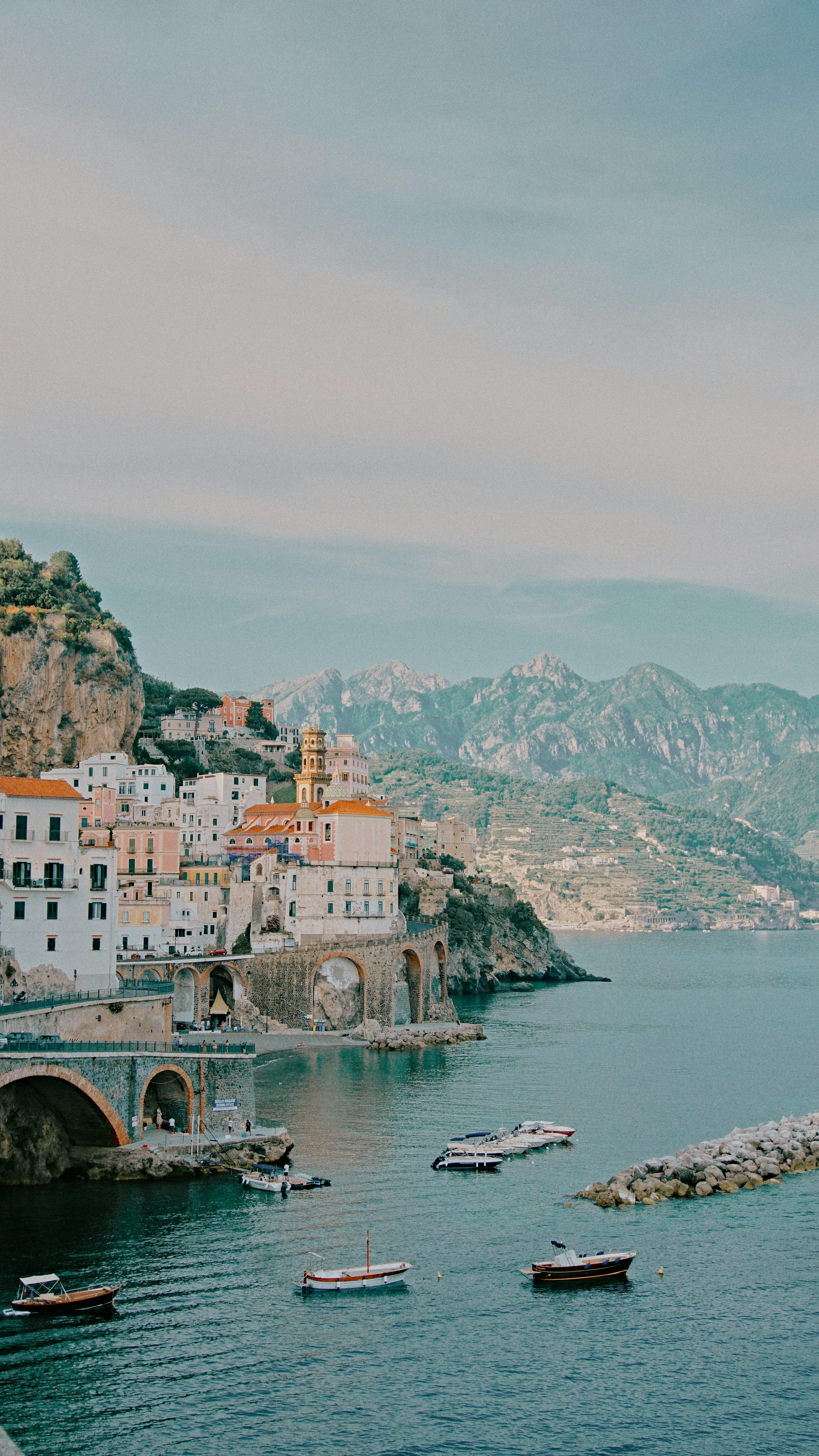 Scenic view of Amalfi Coast with colorful seaside buildings, arched bridge, and boats on turquoise water against mountain backdrop.
