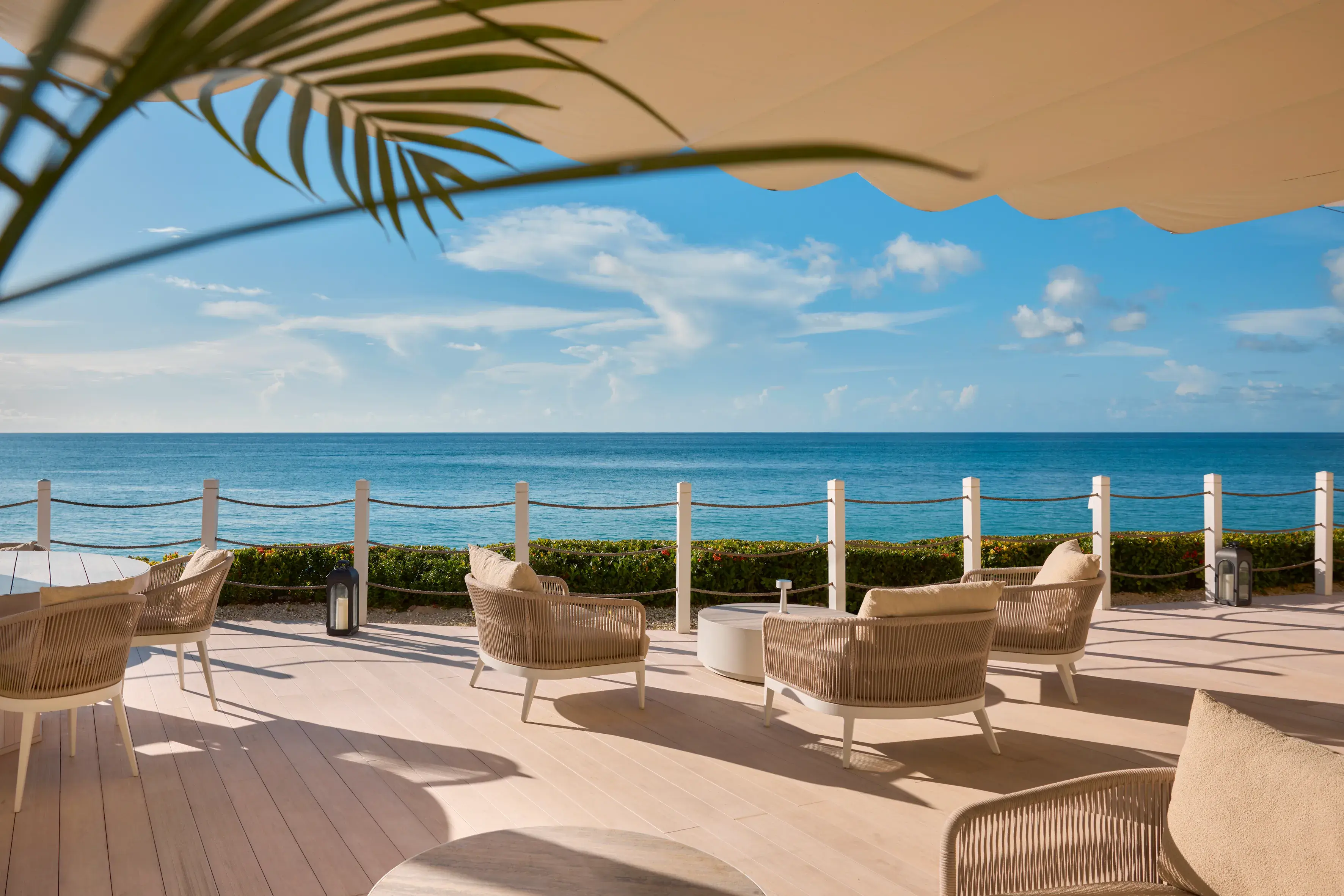 An expansive sun terrace with comfy seating beneath a pale awning overlooking a calm ocean under a clear sky