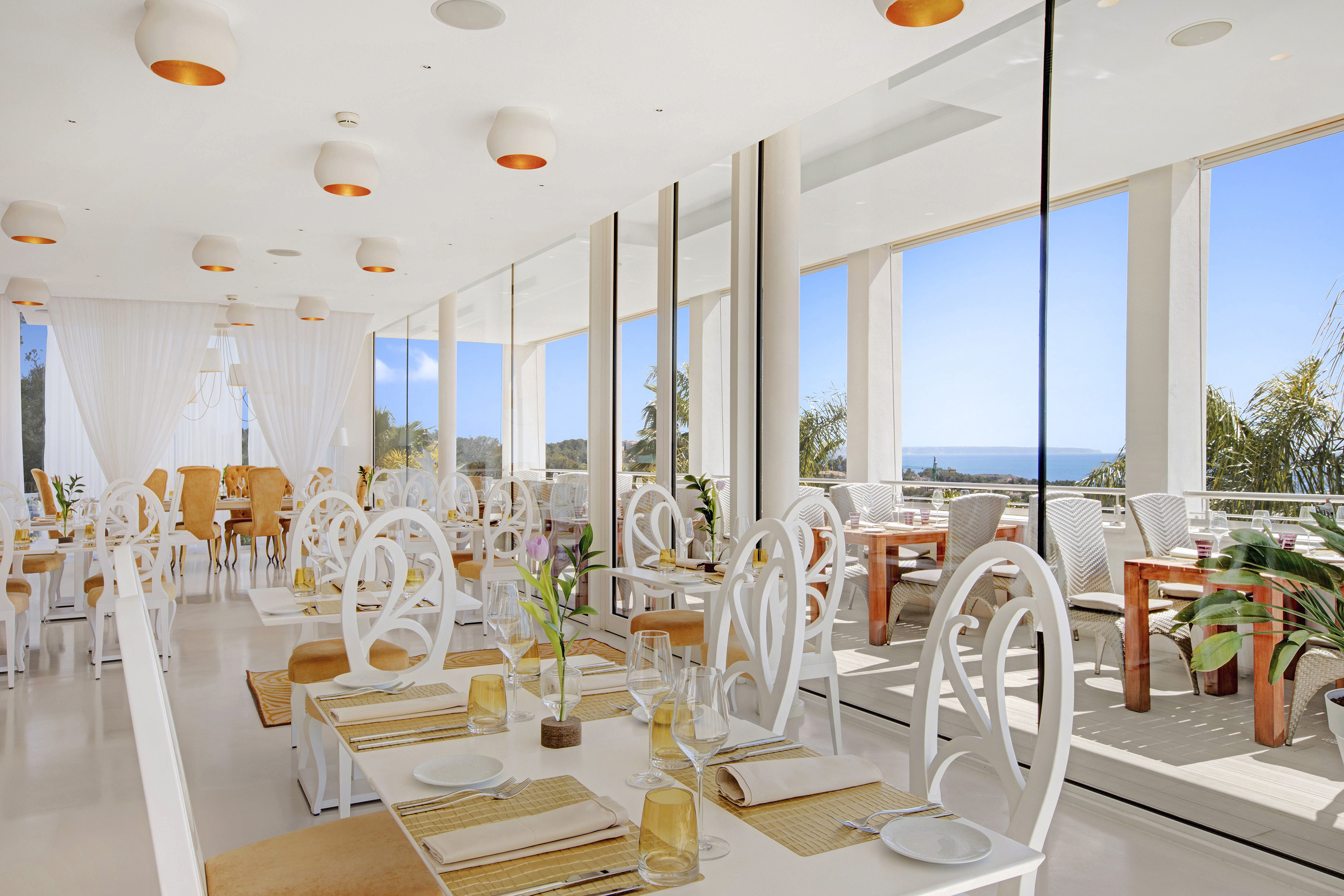 Elegant white dining area at Collins Restaurant at Portals Hills in Mallorca with sea views through large windows and neatly arranged tables.