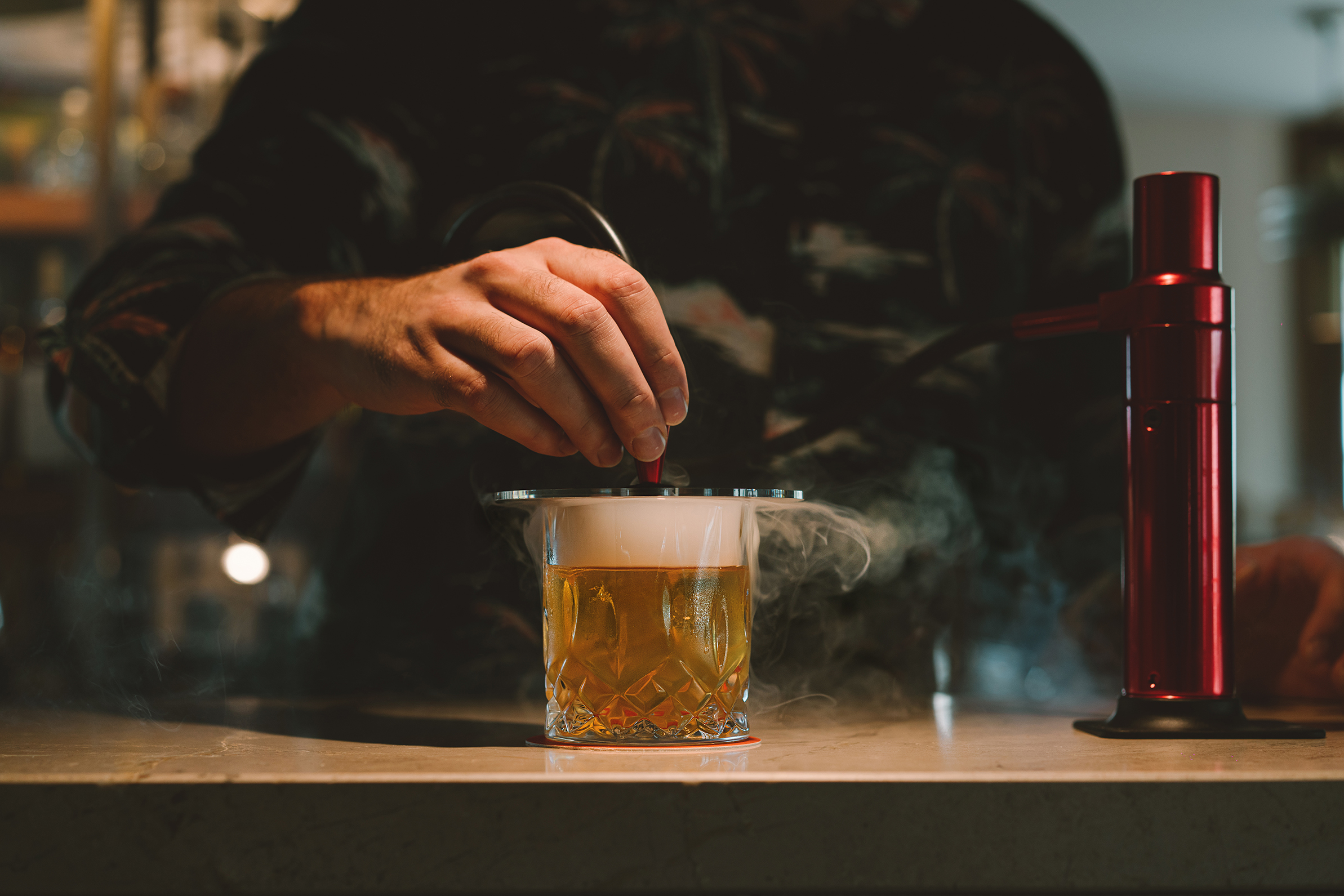 A person preparing a cocktail with steam rising from the glass, next to a red handheld smoking device on a bar counter