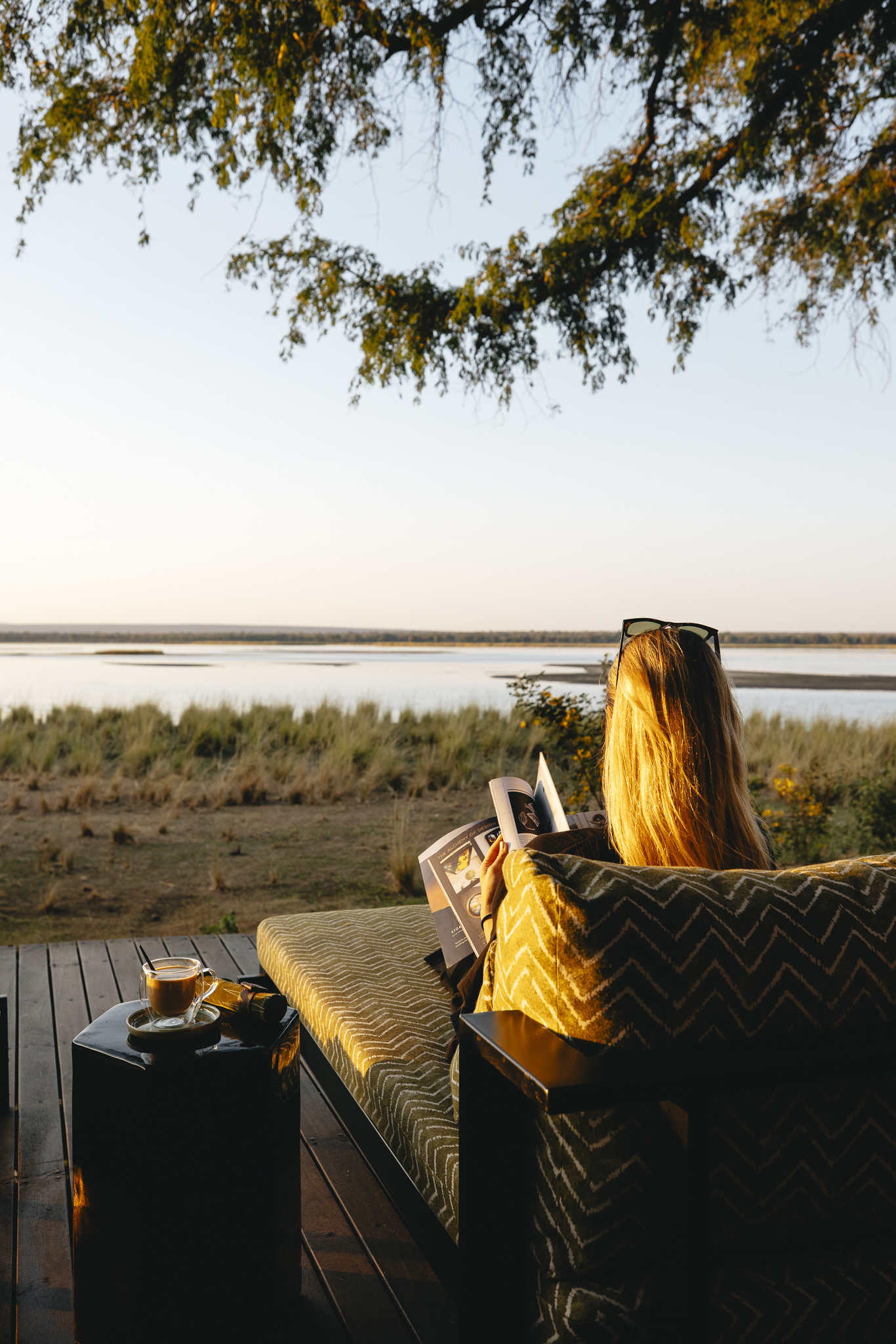 A woman flipping through a magazine with a coffee beside her while sat on a deck overlooking the Zambezi River