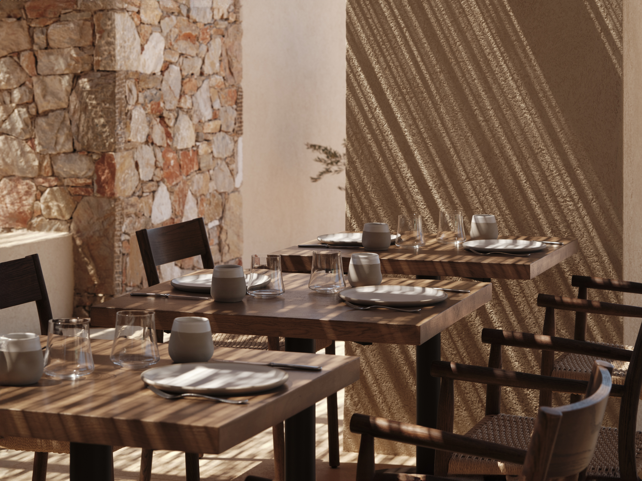 Stylish restaurant dining tables set for dinner set against a stone wall with light streaming in through a woven rooftop